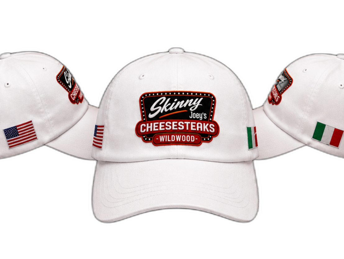Three white baseball caps shown from the front, with the center cap facing forward and the side caps angled inward. The center cap features a large embroidered patch reading 'Ginny 2023 CHEESESTERKS WILWOOD' with a red and black design, and the side caps have small embroidered patches including an American flag and the Italian flag.