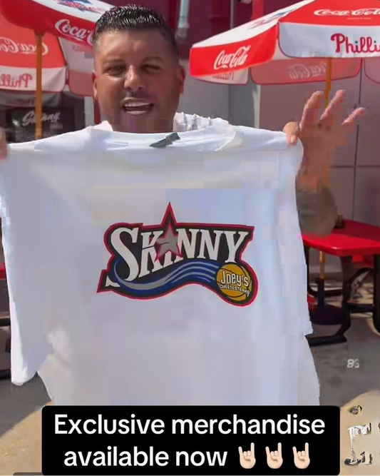 A man outdoors holds up a white t-shirt featuring a large 'Skinny Judo' logo on the front. The background shows red and white Coca-Cola branded umbrellas and tables, with a text overlay at the bottom that reads 'Exclusive merchandise available now'.