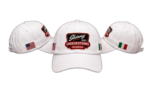 Three white baseball caps shown from the front, with the center cap facing forward and the side caps angled inward. The center cap features a large embroidered patch reading 'Ginny 2023 CHEESESTERKS WILWOOD' with a red and black design, and the side caps have small embroidered patches including an American flag and the Italian flag.