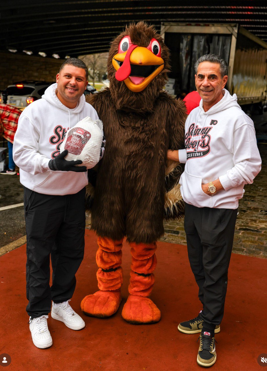 Two men in white hoodies posing with a turkey mascot on a red surface.