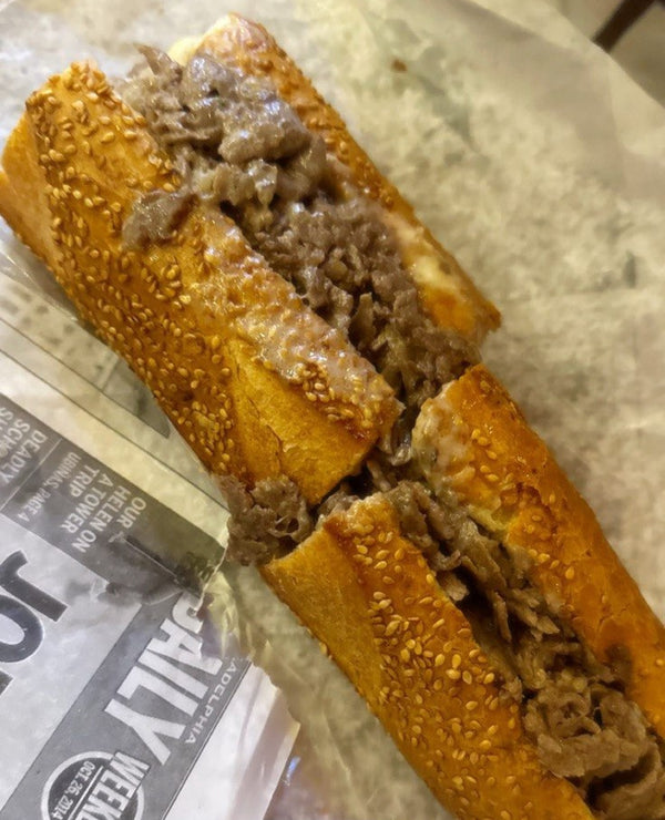 A close-up of a cheesesteak sandwich on a sesame seed baguette, cut open to show the meat filling. The sandwich is placed on paper wrapping with a printed social media overlay that reads 'HAPPY NATIONAL CHEESESTEAK DAY !!' and '@SKINNYJOEYS_CHEESESTEAKS'.